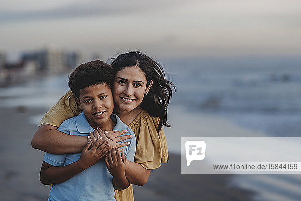 Nahaufnahme einer Mutter  die einen Jungen im Schulalter am Strand lächelnd umarmt