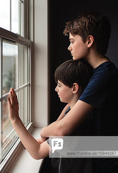 Two boys looking out of a window together with sad faces.