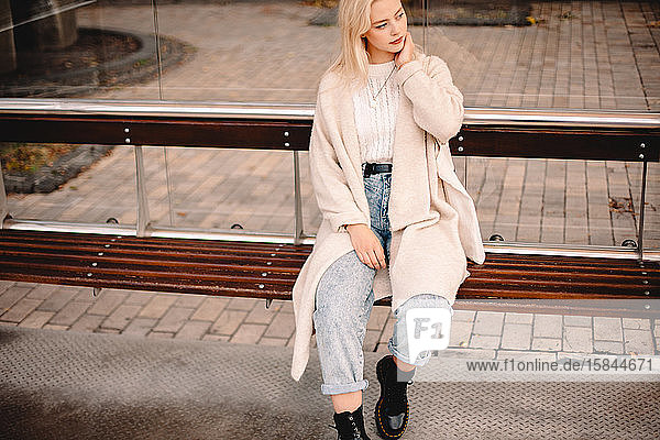 Nachdenkliche stilvolle junge Frau sitzt an Bushaltestelle in der Stadt
