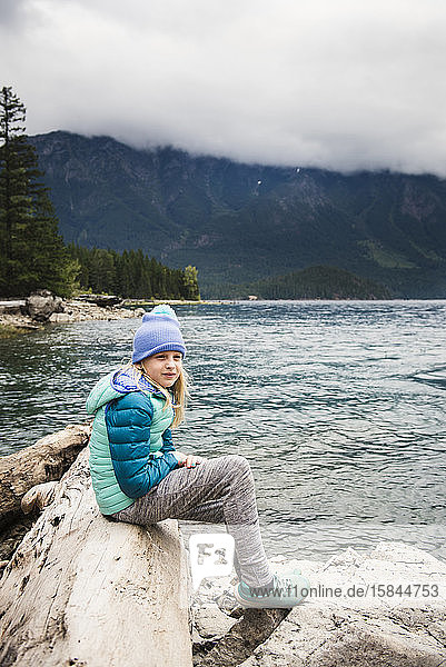 Young Girl Sitting on Log Against Lake and Mountains