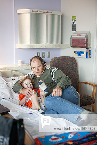 Besorgter Vater kuschelt mit albernem Kleinkind im Krankenhausbett.