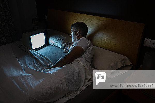 Ein Mann sieht nachts im Dunkeln auf dem Bett einen alten Fernseher.