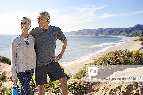 Smiling senior couple standing on cliff at beach against sky during sunny day