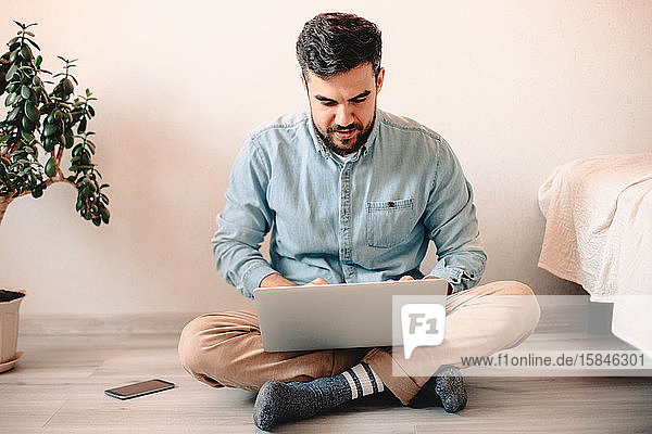Junger Mann benutzt Laptop-Computer  während er zu Hause auf dem Boden sitzt