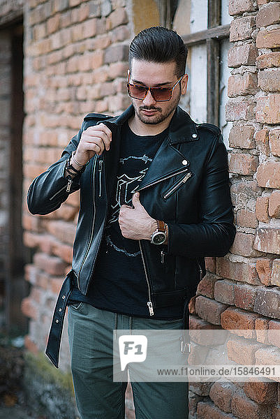 Hipster Man with sunglasses in a jacket standing by a brick wall.