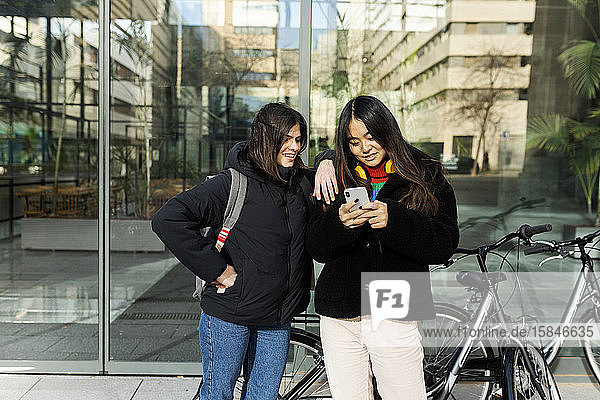 Zwei Freunde schauen auf das Smartphone das in der Glaswand des Gebäudes steht