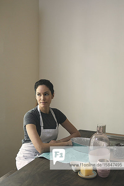 Nachdenkliche Frau in Schürze sitzt mit Behältern und Zutaten für die Zubereitung von Speisen zu Hause