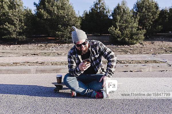 Moderner junger Mann sitzt auf einem Longboard und schaut auf sein Mobiltelefon