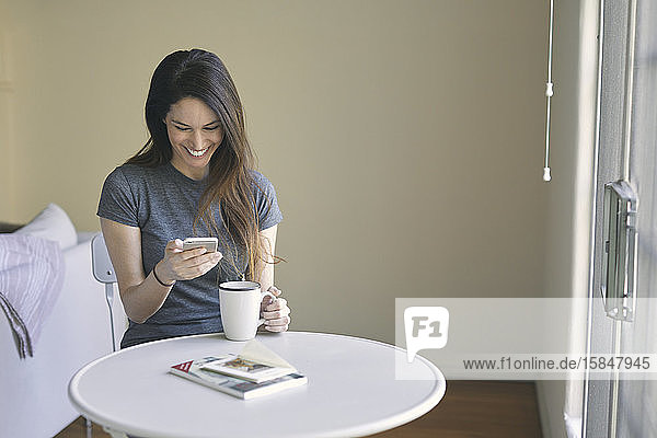 Glückliche Frau benutzt Mobiltelefon  während sie im Wohnzimmer Kaffee am Tisch an der Wand trinkt