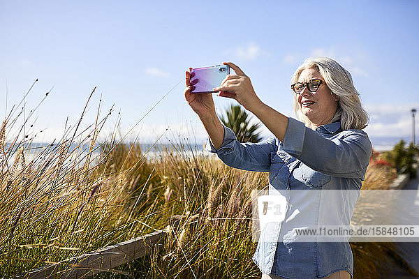 Ältere Frau fotografiert mit Smartphone  während sie am Strand von Manhattan gegen den Himmel steht