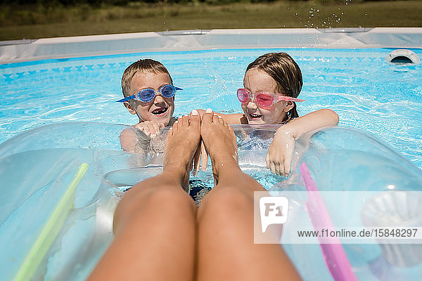Mutter schwimmt auf einem Floß im Pool  während die Kinder an einem sonnigen Tag zu Füßen planschen