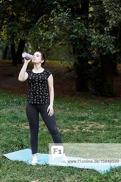Das entspannte Mädchen macht im Park auf einem Teppich Yoga und trinkt w