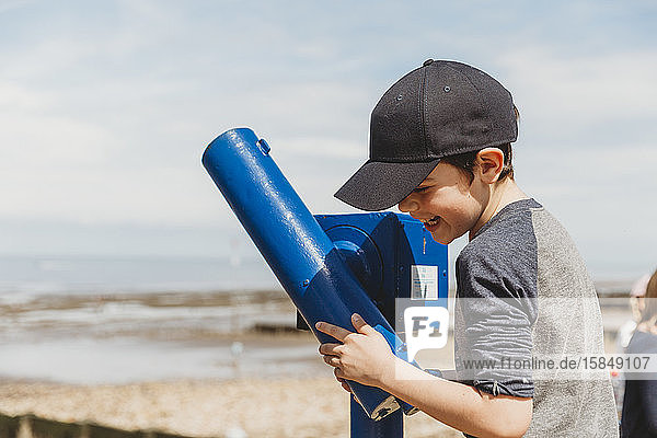 Boy in cap laughing standing next to telescope at seaside