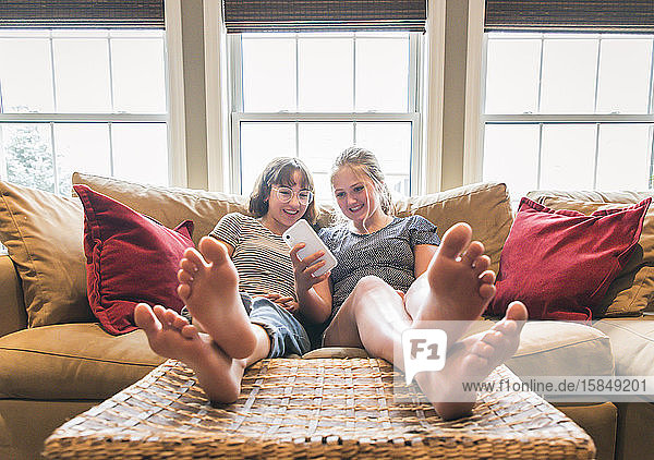 Two teenage girls sitting on couch with feet up looking at cellphone.