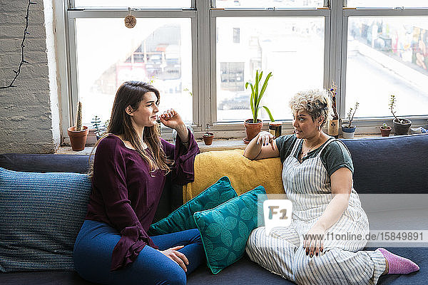 Female roommates talking while relaxing together on sofa against window at home
