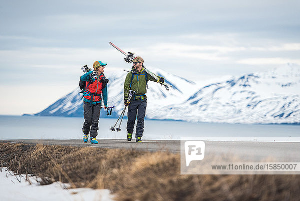 Ein Ehepaar geht in Island mit Skiausrüstung eine Straße entlang