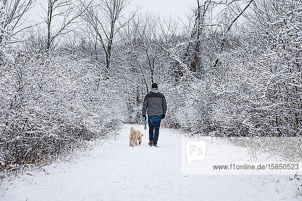Mann  der an einem Wintertag mit einem Hund auf einem verschneiten Pfad durch den Wald spaziert.