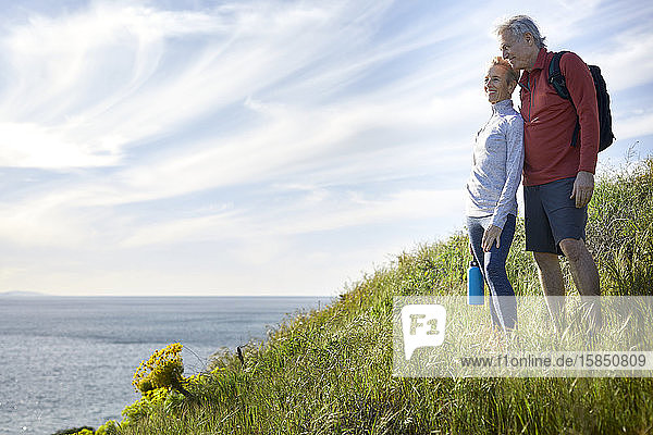 Lächelndes Paar schaut aufs Meer  während es auf einer Klippe gegen den Himmel steht