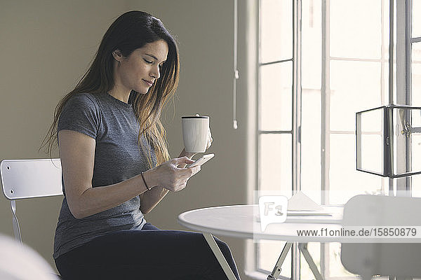 Woman using mobile phone while having coffee at table by window in living room