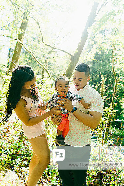 A family of three with a baby boy stand together on a summer day