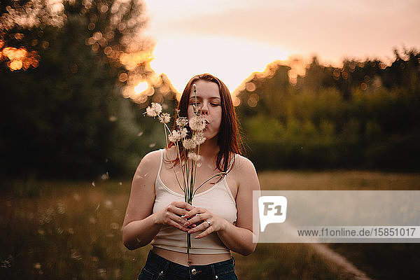 Young carefree woman blowing bunch of dandelions on field in forest