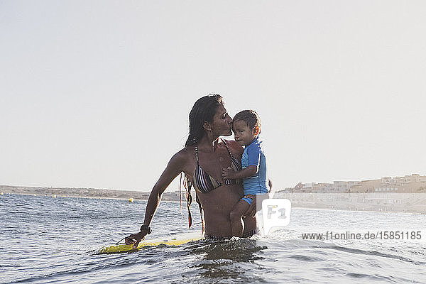 Portrait of a surfer mother holding her son next to a surfboard in sea