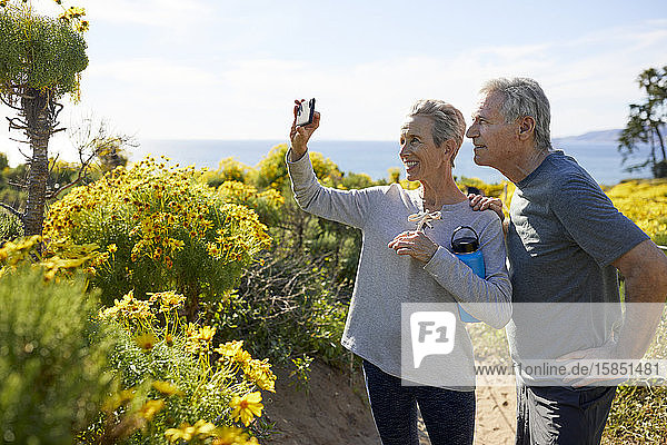 Lächelndes älteres Paar  das sich per Smartphone selbstständig macht  während es am sonnigen Tag auf einer Landschaft am Meer gegen den Himmel steht