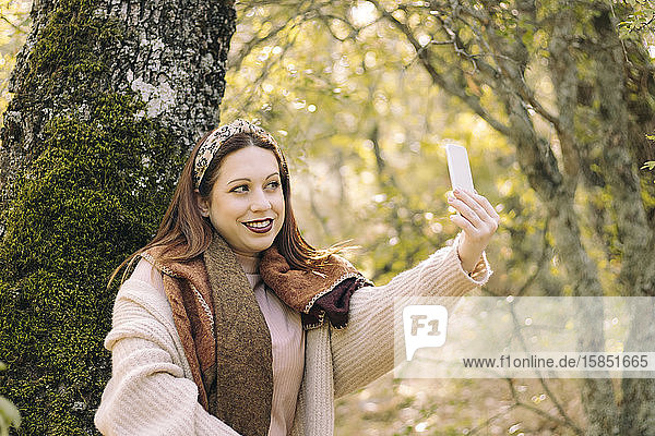 Lächelnde junge Frau  an einen Baum gelehnt  mit ihrem Mobiltelefon