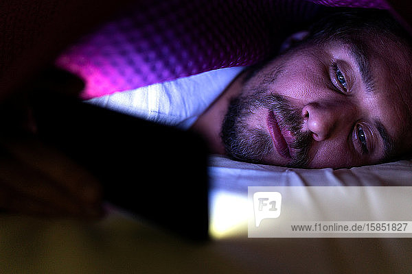 Bearded young man is lying in bed under his blanket looking at phone