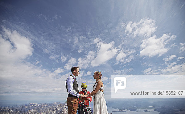 Braut und Bräutigam halten Händchen und heiraten auf einem Berggipfel mit blauem Himmel
