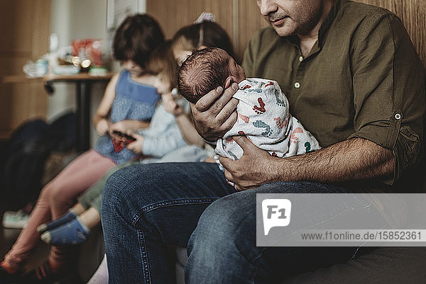 Side view of father holding newborn with siblings in background