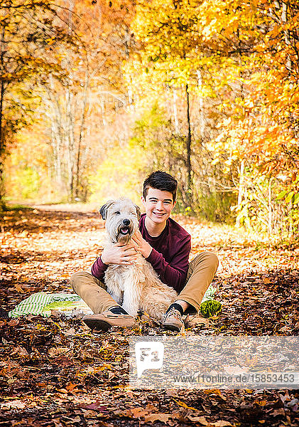 Teenage boy sitting with dog on leafy trail in the woods on fall day.