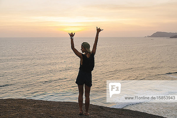 Junge Frau steht bei Sonnenuntergang auf einem Felsen am Strand