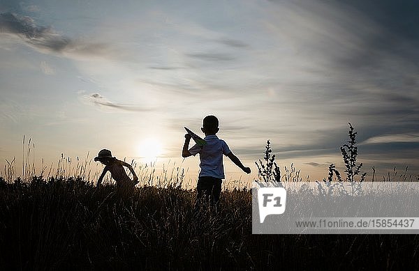 Silhouette von Geschwistern  die bei Sonnenuntergang draußen mit Papierflugzeugen spielen