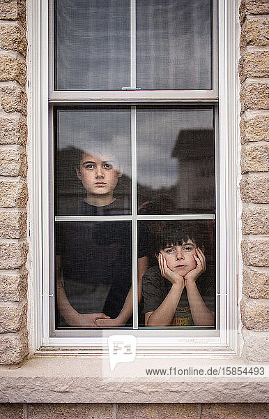 Two boys looking out of a window together with bored faces.