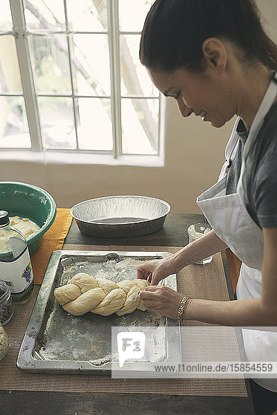 Smiling young woman preparing Challah bread from dough in tray on table at home