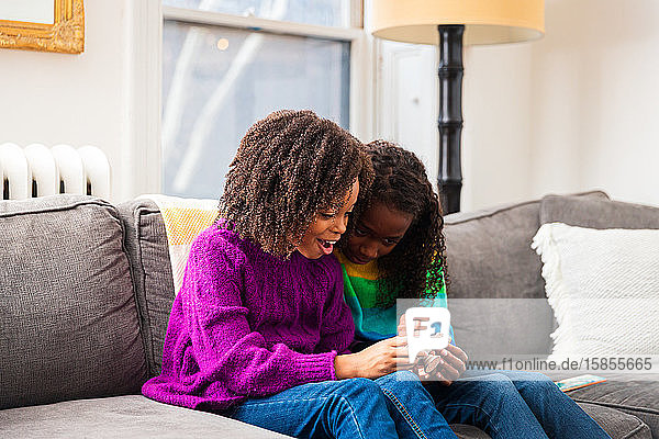 Glückliches Mädchen zeigt seiner Schwester ein Smartphone  während sie im Wohnzimmer auf dem Sofa sitzt