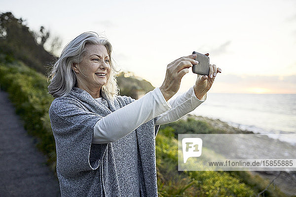 Seitenansicht einer fröhlichen älteren Frau  die mit einem Smartphone am Manhattan Beach bei Sonnenuntergang fotografiert