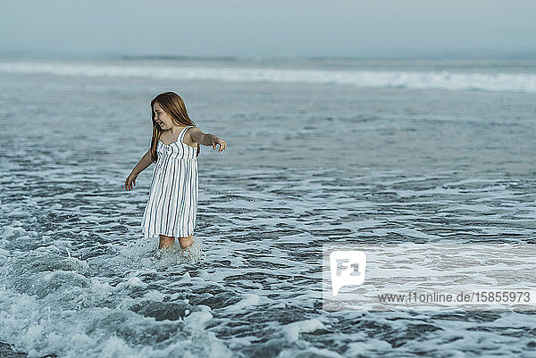 Junges rothaariges Mädchen im Kleid spielt in der Abenddämmerung in Meereswellen