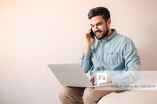 Glücklicher Mann telefoniert mit einem Smartphone  während er zu Hause einen Laptop benutzt