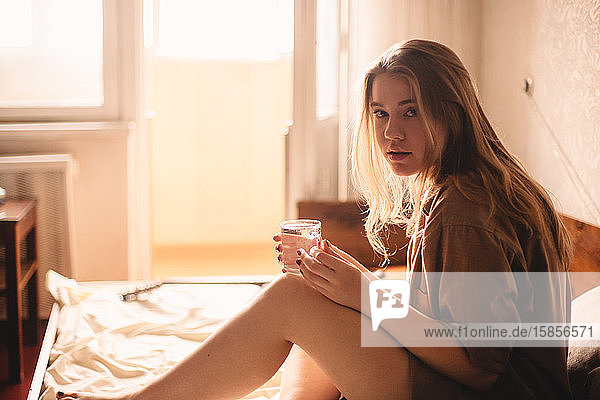 Young woman holding glass of water while sitting in bed at home