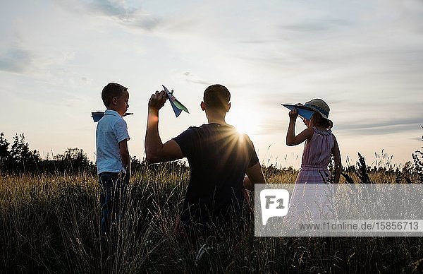 Vater spielt mit Sohn und Tochter auf einer Wiese bei Sonnenuntergang Flugzeuge