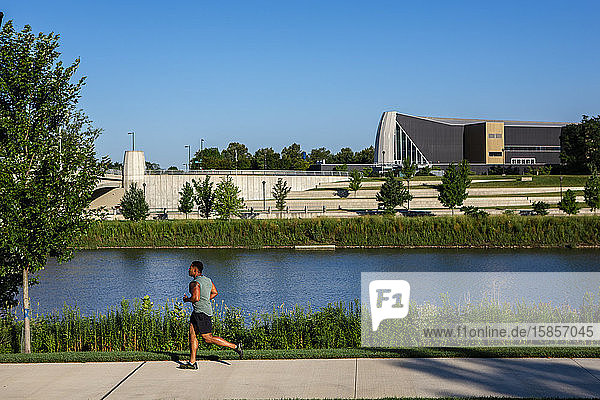 Ein Mann joggt in einem Stadtpark auf einem Weg gegen einen Fluss und hohe Gebäude