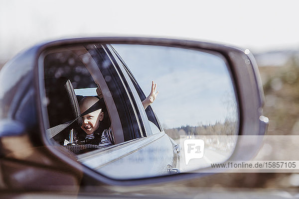 Car mirror shot of boy in back seat with hand out window on sunny day