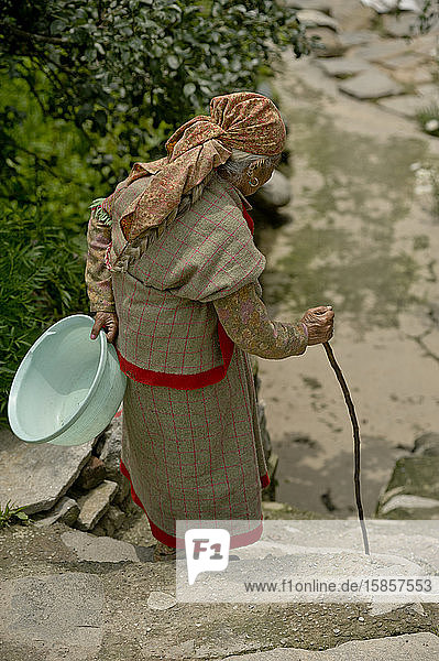 Alte Kullu-Frau geht mit Stock und Plastikbecken eine Treppe hinunter