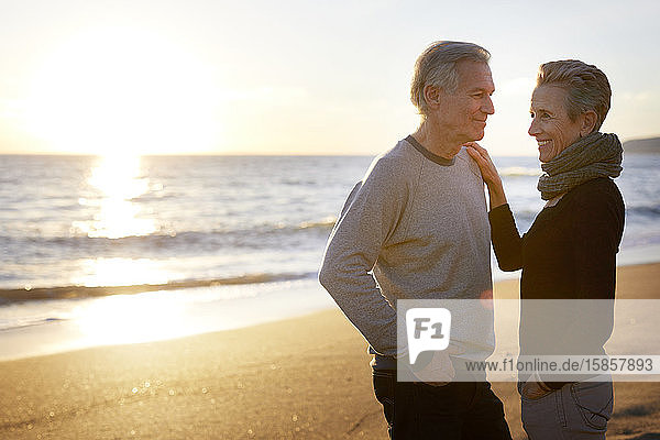 Lächelndes älteres Paar steht sich bei Sonnenuntergang am Strand gegenüber