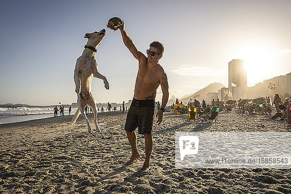 Mann spielt mit springendem Hund und Kokosnuss am Strand von Copacabana