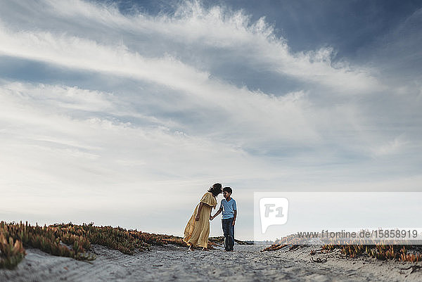 Wide view of mother kissing son on beach against cloudy blue sky