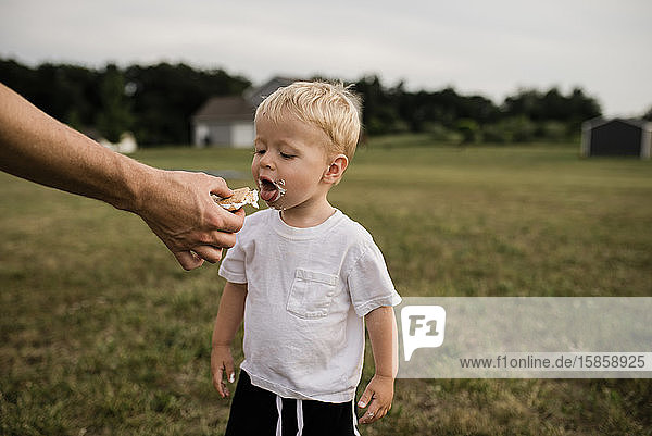 Toddler boy opens mouth to eat s'more from dad's hand in backyard