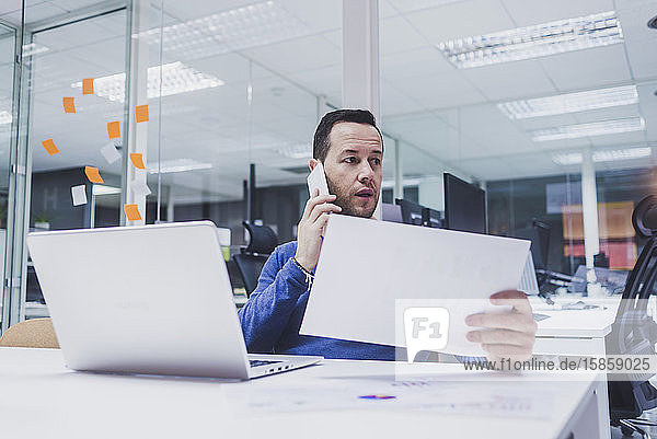 Männlicher leitender Angestellter telefoniert mit einem Mobiltelefon  während er am Schreibtisch einen Laptop benutzt